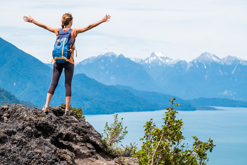 Young woman hiker stands on the rock with stretched hands and enjoys the mountains view