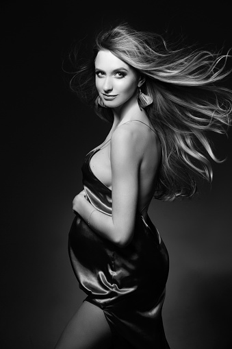 Monochrome portrait of gorgeous and beautiful pregnant woman in sexy silk dress, embracing belly. Fashionable mom posing at dark studio, looking at camera. Concept of pragnancy fashion. Windy effect.