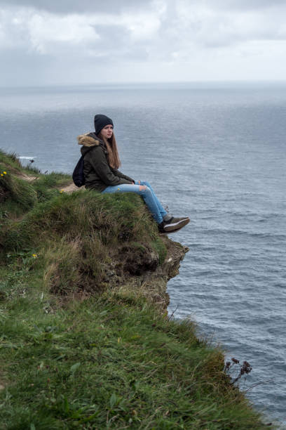mädchen sitzt am rande der steilen klippen von moher in irland - cliff at the edge of grass sea stock-fotos und bilder