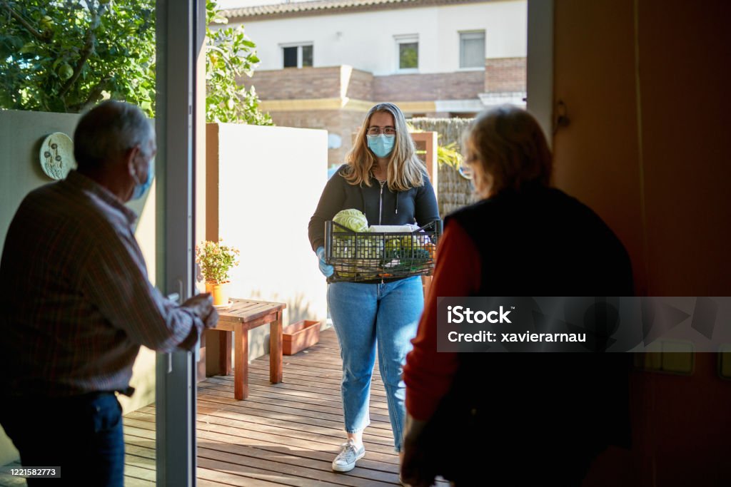 孫娘がパンデミック中に屋外で食料品を配達している間、屋内に立っている祖父母の背面図 - ボランティアのロイヤリティフリーストックフォト