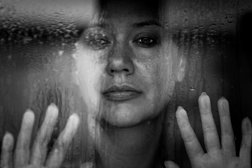 Close up black and white image depicting a sad, depressed-looking woman in her 30s, and of caucasian ethnicity, looking of a rain-soaked window with her hands pressed to the glass. The image was taken during the Covid-19 pandemic during lockdown, and the image illustrates a mental health concept, as well as a domestic abuse concept. Room for copy space.