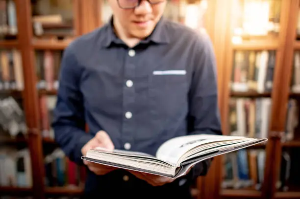 Smart Asian man university student wearing glasses reading book by vintage bookshelf. Textbook resources in college library for educational subject and research. Scholarship for education opportunity.