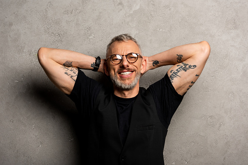 happy and tattooed man in glasses looking at camera on grey