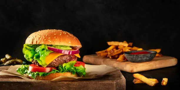 Photo of A panorama of a burger with beef, cheese, onion, tomato, and green salad, a side view on a dark background with pickles and French fries, with a place for text