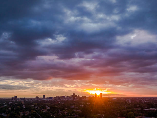 The autumn/summer sun creates sunrays between two tall buildings as it sets beneath dark, moody skies over the Adelaide Central Business District seen in the distance from the leafy eastern suburbs.
