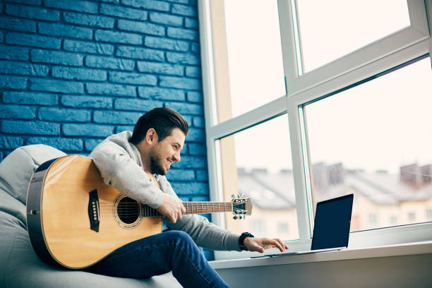 happy guitarist with instrument sitting at modern apartment and using laptop stock photo