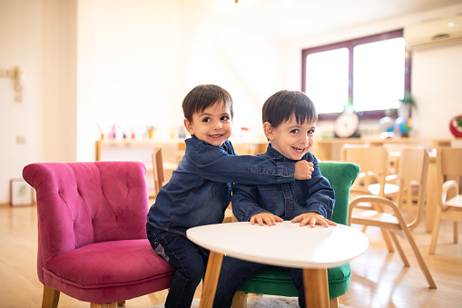 Cute twin brothers sitting on small wooden chair and, embracing and having fun time together in child care classroom, smiling and looking at camera