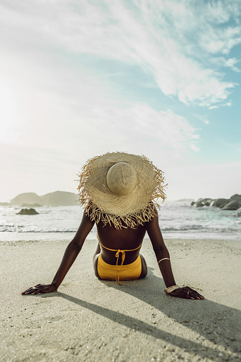 Rear view of a woman relaxing on beach. African female model in bikini wearing a straw hat on sea shore. Woman on beach vacation.