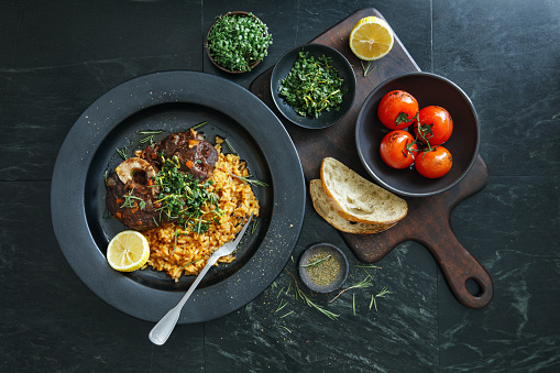 Traditional Italian Osso Buco with Creamy Saffron Risotto dish topped with fresh parsley, garlic and lemon zest.  Flat lay top-down composition on dark background.