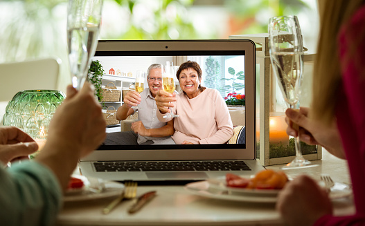 Man and woman sitting at the dining table, having dinner, drinking Champagne and having video call with senior parents on laptop. Staying home, quarantine and social distancing celebration of event.