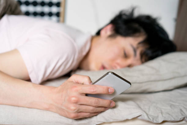 uomo asiatico che si sveglia a letto con allarme smartphone al mattino - dormire troppo foto e immagini stock