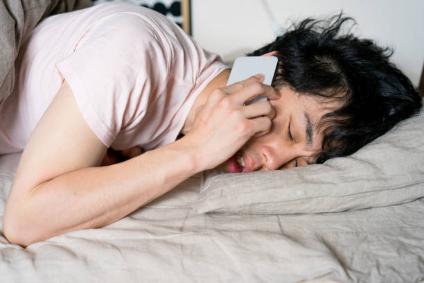 Asian man making a smartphone call from bed in the morning Asian man making a smartphone call from bed in the morning.
He has Bedhead. oversleeping stock pictures, royalty-free photos & images