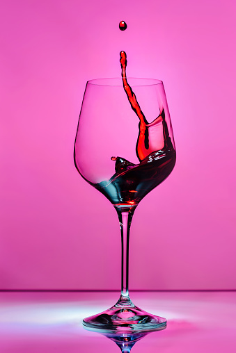 Red wine splashing in a glass on a pink background with blue reflexes and reflection in a glass countertop.