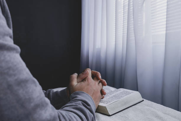 istock Man reading  Holy Bible on a white table, black wall and window  background. Stay at home. Religion concept. 1221647406