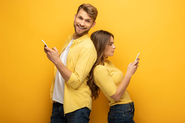 istock Side view of man smiling at camera near girlfriend using smartphone on yellow background 1221651741
