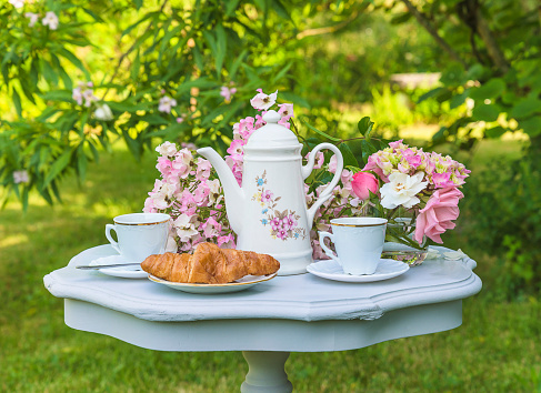 Vintage coffee pot, two cups of coffee, croissant and bunch of rose flowers on vintage table in the garden