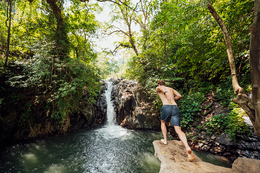 A rear view action shot of a young man taking a running leap off a cliff next to a waterfall in Bali.