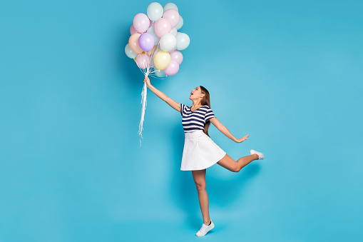 Full size profile photo of beautiful funky lady hold many colorful balloons fly up wind blowing wear striped t-shirt white short skirt footwear isolated blue color background