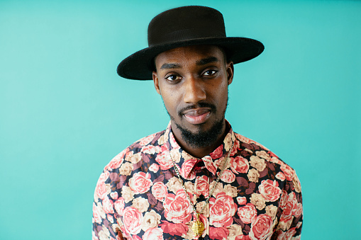 Portrait of a young man in cool flower shirt and black hat against blue studio background