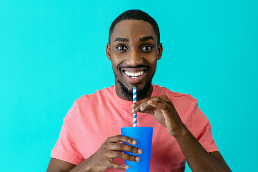 Portrait of a happy young man smiling and drinking with a paper straw from a plastic cup, against blue studio background