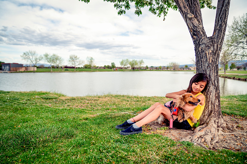 A beautiful mixed race young girl resting by a tree with her dog, cuddling and petting him