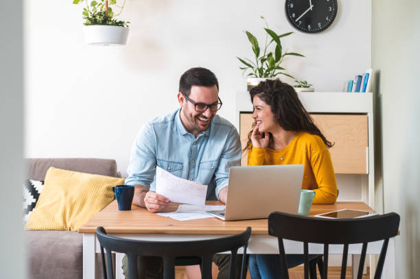 pareja sonriente gestionando las finanzas, revisando sus cuentas bancarias utilizando el ordenador portátil y la calculadora en la foto moderna de la cocina - computer laptop couple women fotografías e imágenes de stock