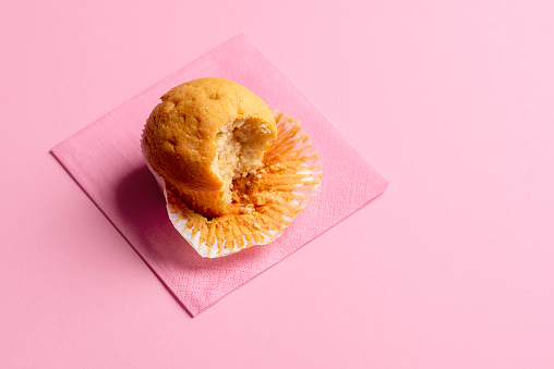 Basic muffin with a missing bite on a napkin, on a seamless pink background. Just one eaten muffin. Partially eaten cupcake. Popular dessert.