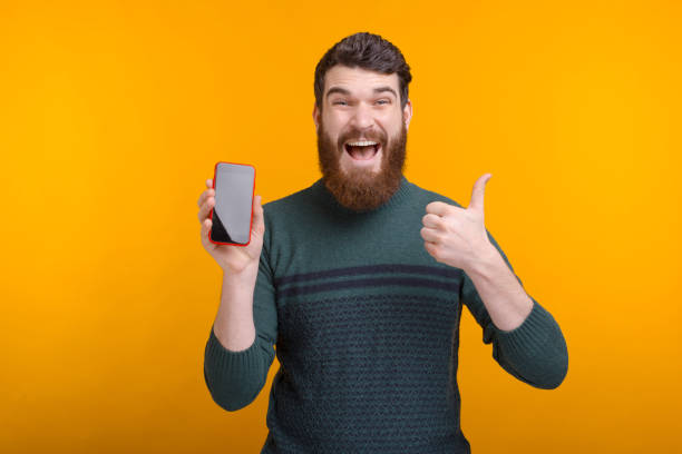 istock Exited bearded man is looking at the camera showing thumb up and his phone screen. 1221684062