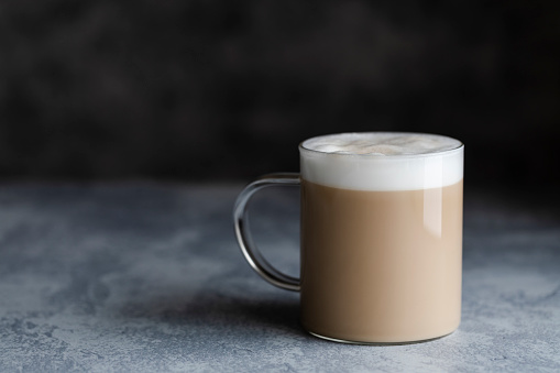 A cup of cafe latte, cafe au lait or chai latte. The photo shows a beige or light brown drink with a white foam on top. The drink is in a glass mug with a handle, and is on a dark gray background. Copy space room for text.