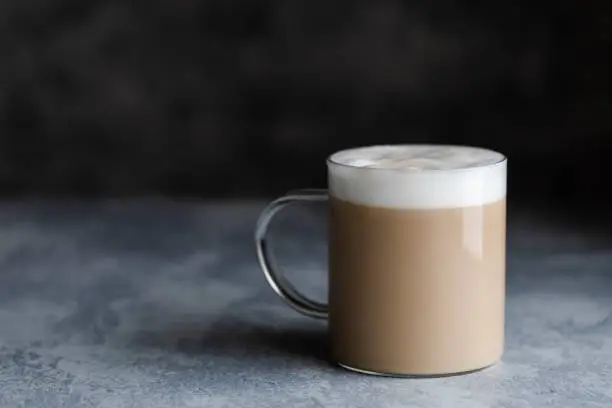 A cup of cafe latte, cafe au lait or chai latte. The photo shows a beige or light brown drink with a white foam on top. The drink is in a glass mug with a handle, and is on a dark gray background. Copy space room for text.