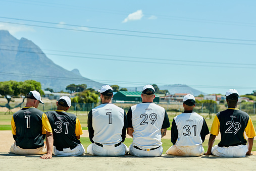 Rearview shot of a team of unrecognizable baseball players sitting together near a baseball field during the day