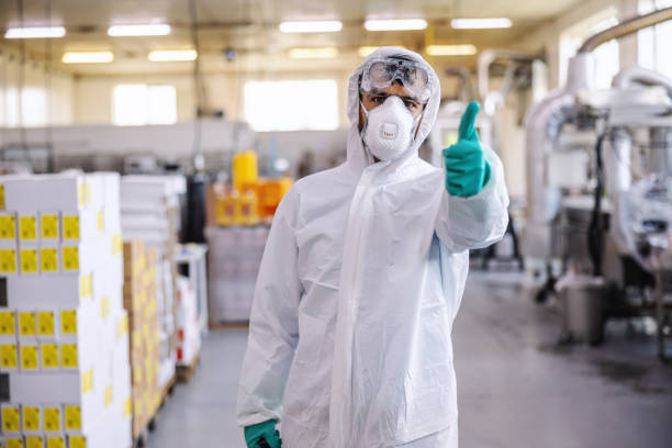 homme dans la combinaison protectrice, le masque et les gants restant dans l’usine de production de nourriture et affichant des pouces vers le haut. il vient de désinfecter toute l’installation du virus covid-19 / corona. - safety sign protective workwear factory photos et images de collection