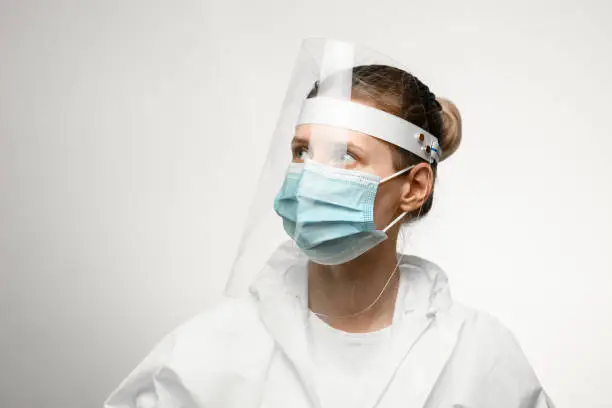 young woman in medical mask and white protective suit with protective shield on her head looking away on grey background.