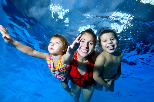 Happy little children, a boy and a girl, together with their mother swim and play under the water in the pool. They smile provocatively and pose for the camera on a blue background. Digital photo