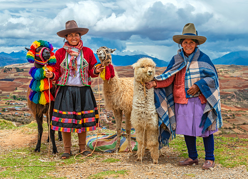 A rural portrait of Quechua Indigenous Women in traditional clothes with their domestic animals, two llama and one alpaca, Sacred Valley of the Inca, Cusco, Peru.