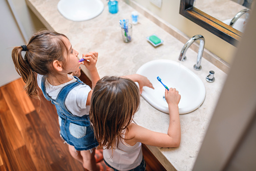 Elevated close-up of 6 and 8 year old sisters standing together at bathroom sink and brushing their teeth.