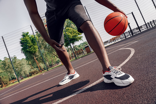 Young african man playing on court outdoors dribbling basketball between legs technique close-up