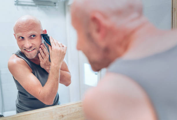 istock Hairless man fixing his beard looking in bathroom mirror using an electric rechargeable Beard Trimmer. Everyday body and skincare routine treatments concept image. 1221714428