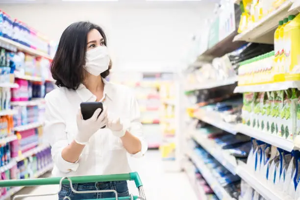 Asian woman wearing face mask and rubber glove push shopping cart in supermarket department store. Girl hold smartphone choose & look grocery things to buy during coronavirus crisis, covid19 outbreak.
