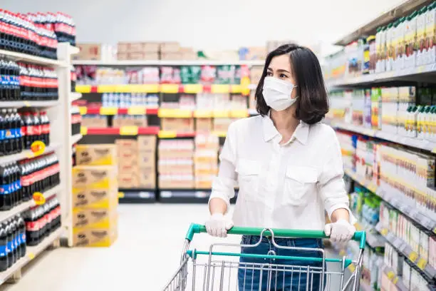 Asian woman wearing face mask and rubber glove push shopping cart in supermarket departmentstore. Girl choosing, looking grocery things to buy at shelf during coronavirus crisis or covid19 outbreak.