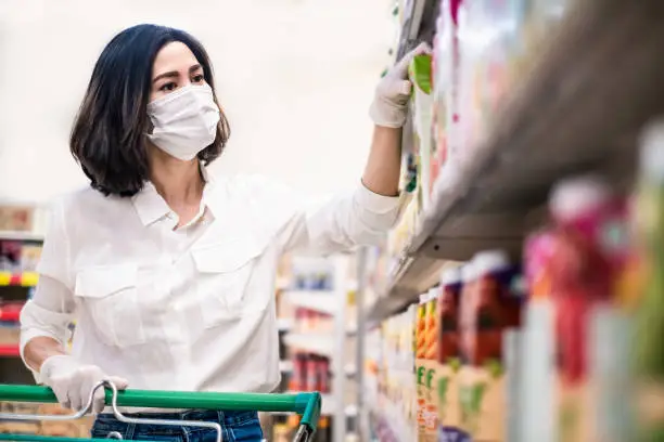 Asian woman wearing face mask and rubber glove push shopping cart in supermarket department store. Girl choosing, looking grocery things to buy at shelf during coronavirus crisis or covid19 outbreak.