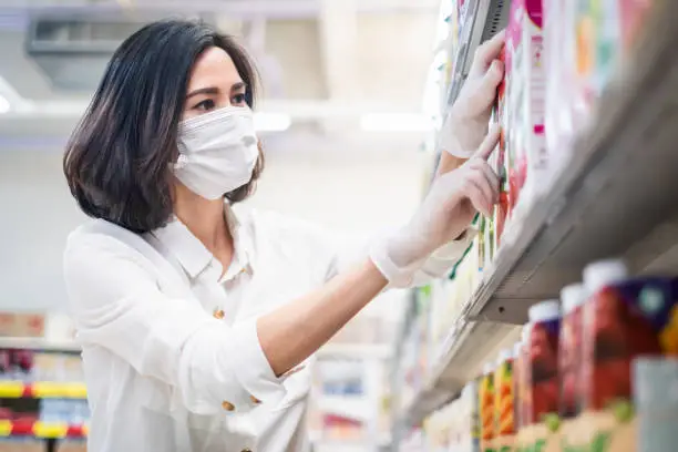 Asian woman wearing face mask and rubber glove shopping beverages in supermarket department store. Girl choosing, looking grocery things to buy at shelf during coronavirus crisis or covid19 outbreak.