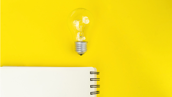 Flat lay of light bulb and empty notebook with yellow background