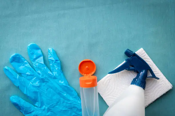 Still life flat lay arrangement of cleaning spray and paper towel, bottle of hand sanitizer, and surgical glove on solid blue background with copy space