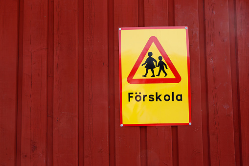 Yellow sign with the beware of children traffic sign and text in Swedish for kindergarten on a red wooden wall.