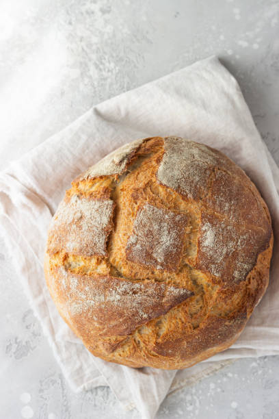 Whole artisan rustic bread from wheat and rye flour on a linen napkin, light grey stone background. Selective focus.