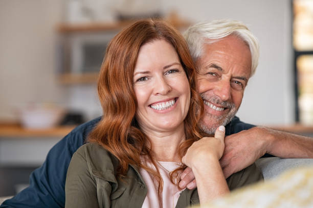 Happy smiling mature couple at home Portrait of middle aged couple hugging and looking at camera. Close up face of happy mature couple having fun at home. Senior man embracing his beautiful wife on the couch while looking at camera together. mid adult couple stock pictures, royalty-free photos & images