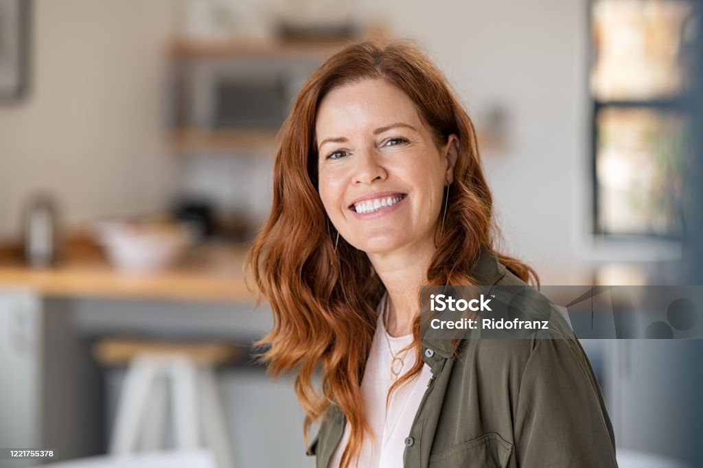 Mature beautiful woman with red hair Portrait of smiling mature woman looking at camera with big grin. Successful middle aged woman at home smiling. Beautiful mid adult lady in casual with long red hair enjoying whitening teeth treatment. Women Stock Photo