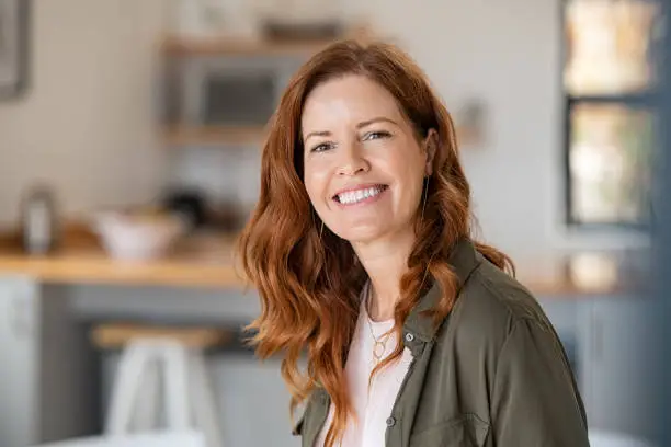Portrait of smiling mature woman looking at camera with big grin. Successful middle aged woman at home smiling. Beautiful mid adult lady in casual with long red hair enjoying whitening teeth treatment.