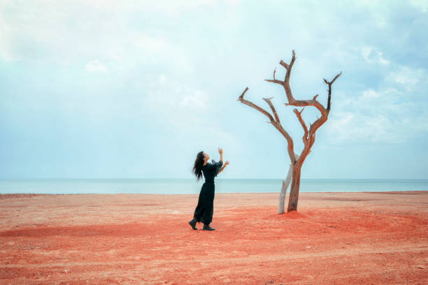 istock Woman in black dress standing on sand with dead tree 1221755397
